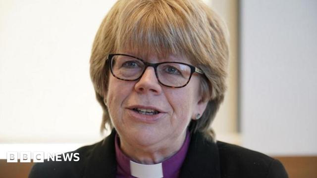 Dame Sarah Mullally is dressed in a dark jacket and is wearing a white clerical collar on a purple shirt. 