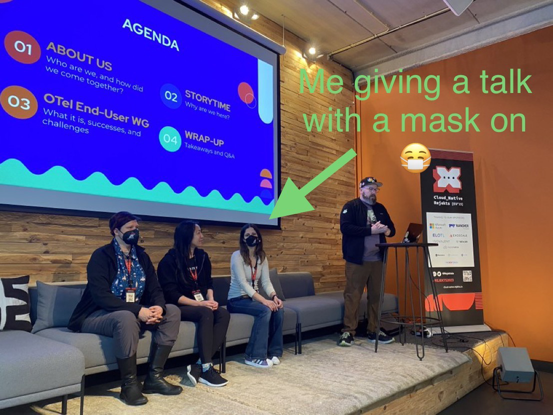 Me sitting on a couch at a conference, surrounded by other speakers, wearing a mask.