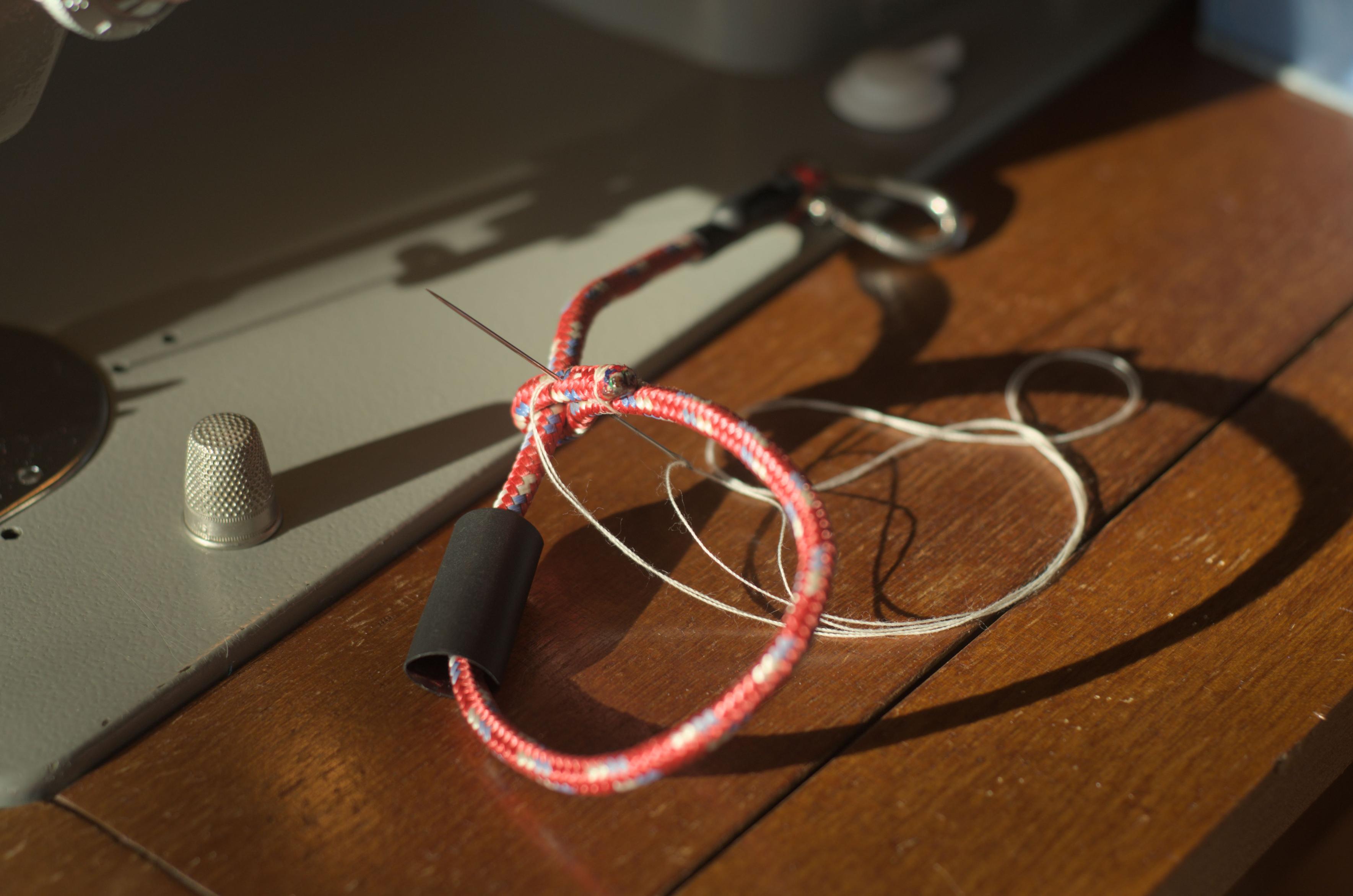 Sewing the melted cut end of the rope around the standing end and into a small loop to create the wrist loop. A long needle with a sturdy white thread sticks through two layers of rope. The black heat shrink tube is already on the loop, to be moved on the sewn section later.