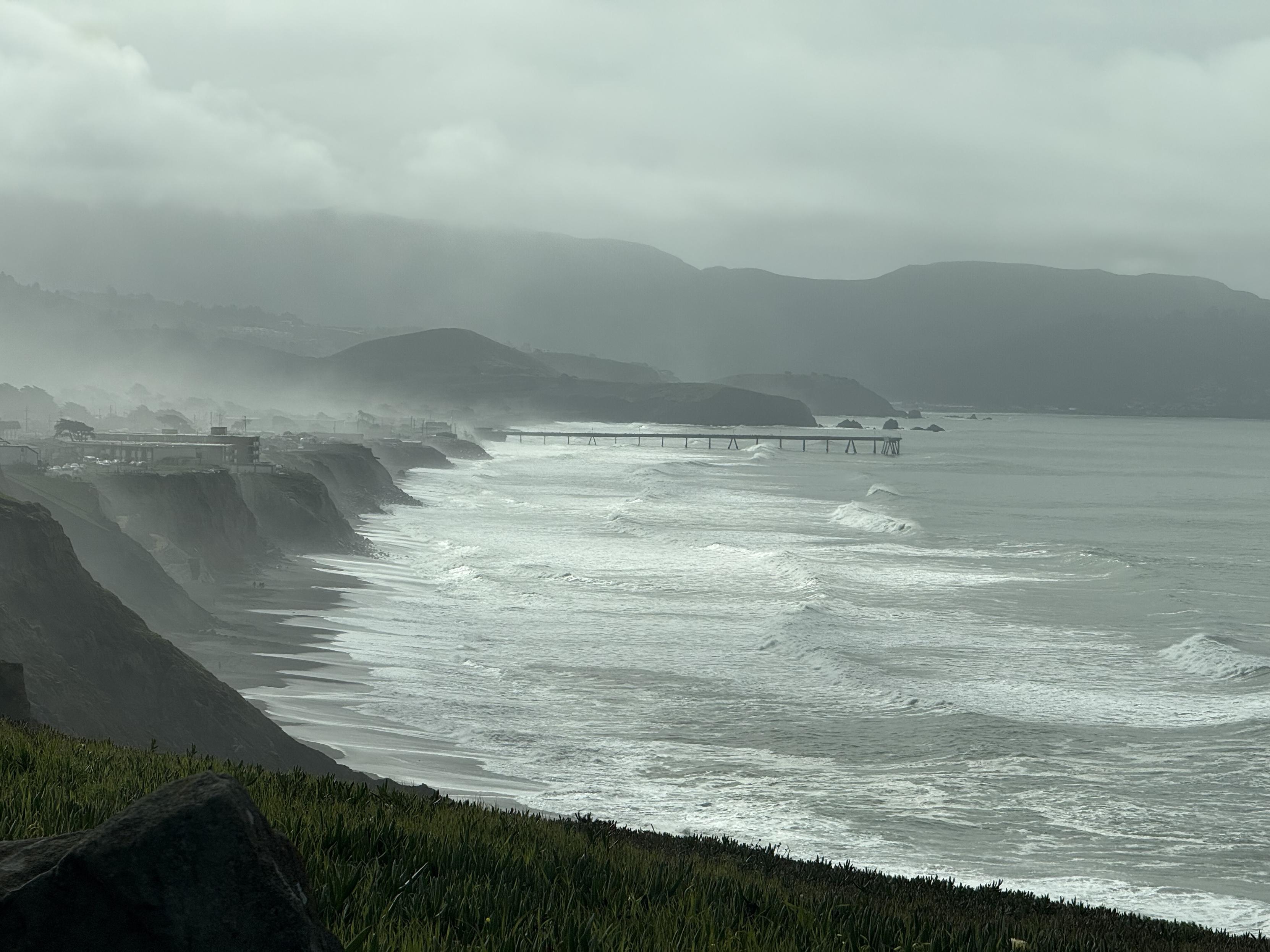 A picture of a coastline with waves rolling against a beach set below cliffs.  In the distance: a pier extends across a third of the frame.  A bit of fog hangs over the land.