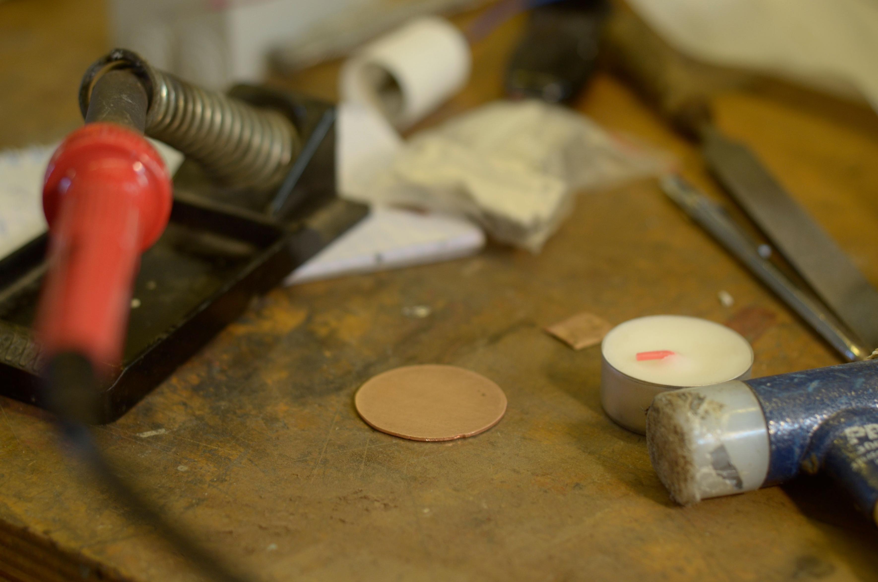 A copper disc in a mess of tools and next to a small candle that has the same radius.