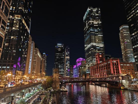 Buildings at night. Red, pink, and white lights reflect from the water below.