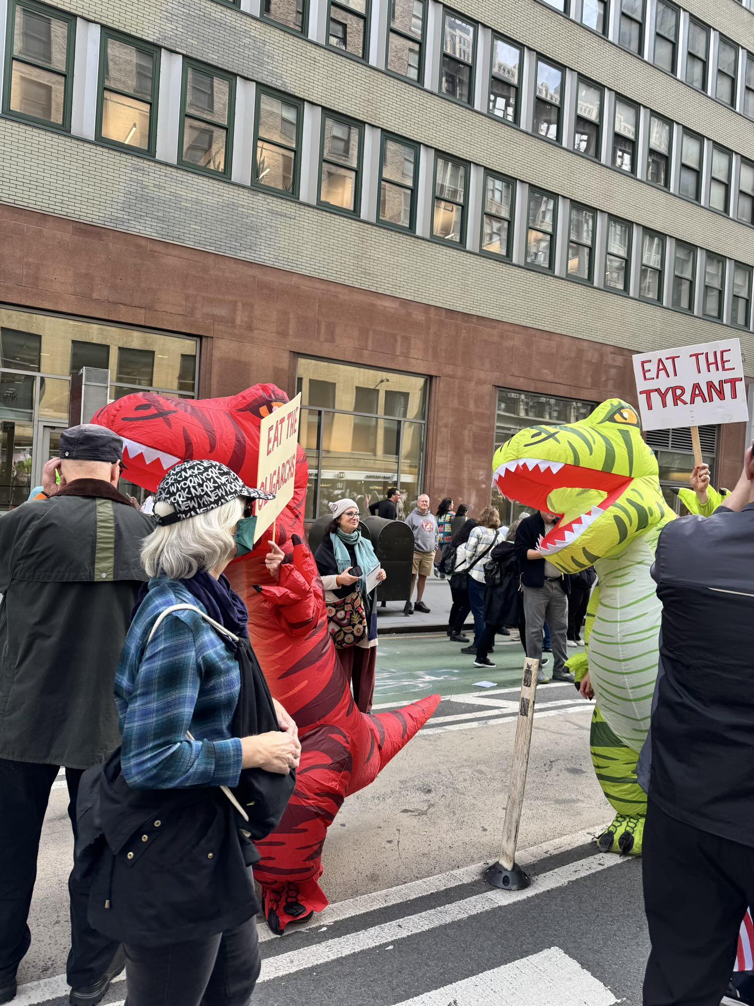 Photo of two people in inflatable T-Rex outfits one of whom is featuring a sign “Eat The Tyrant”