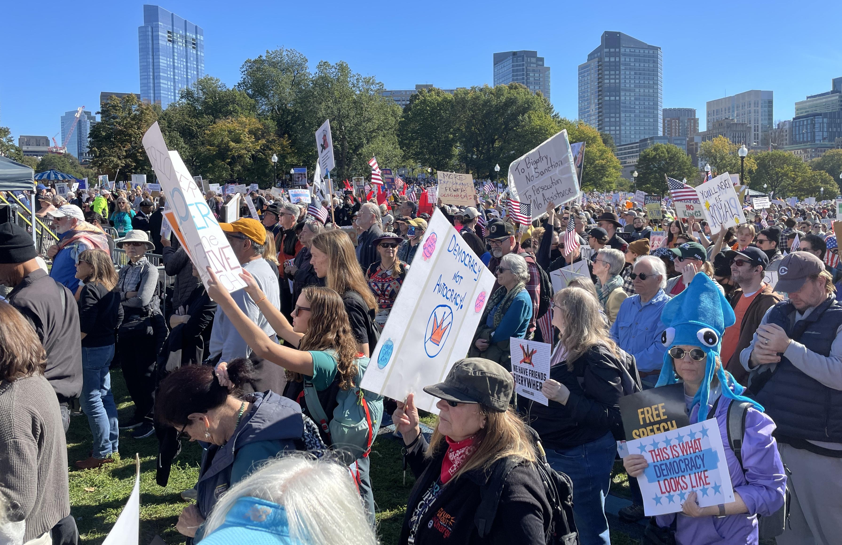 A crowd of people, most of them holding protest signs for 'no kings'. The crowd extends into the distance, where you can see trees and Boston skyline behind them, below a crystal-clear blue sky. Some people are wearing short sleeves, but most have some kind of light outerwear.