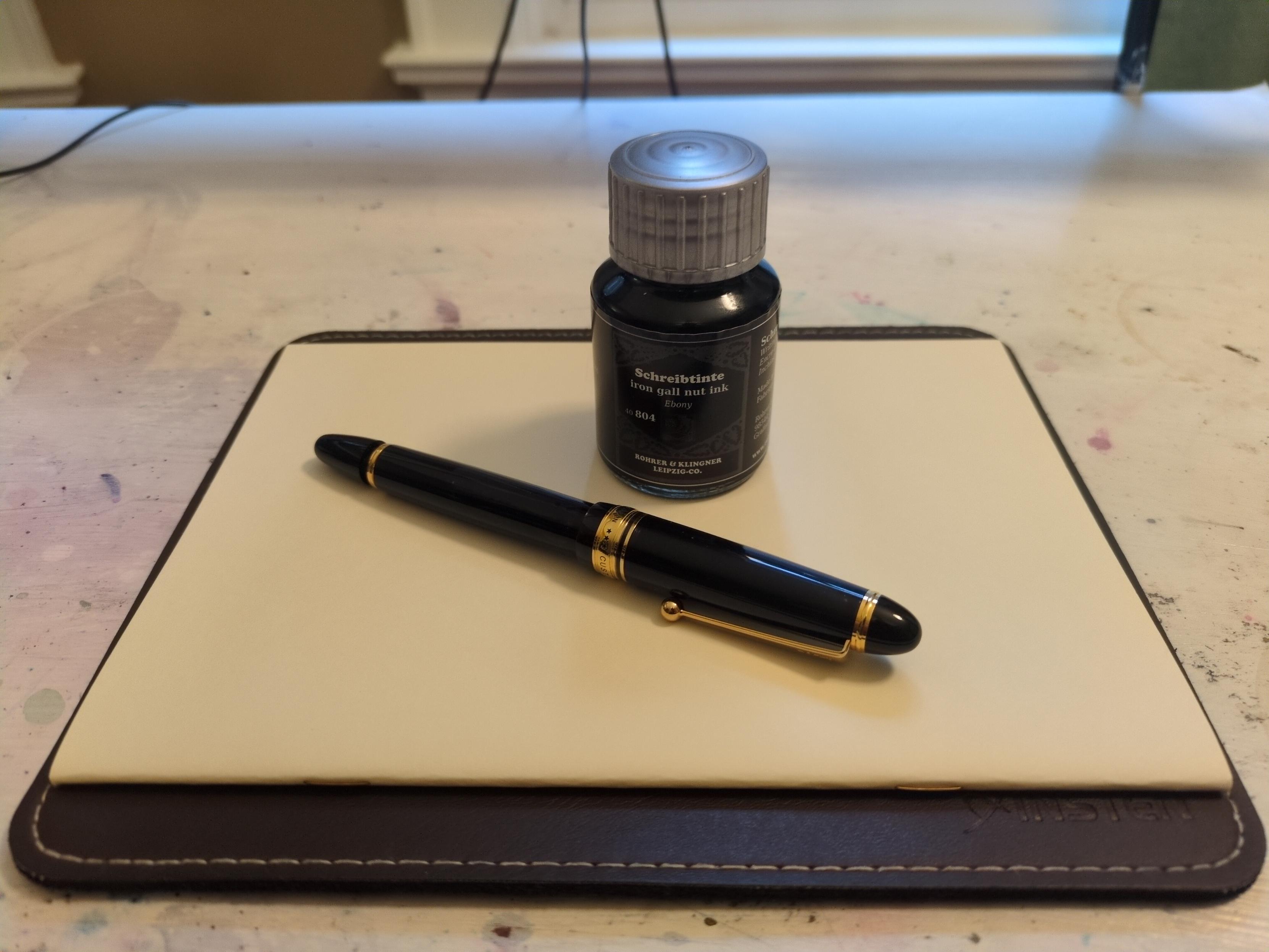A black fountain pen with gold trim and a “cigar” design that gradually tapers to rounded tips. There is a black bottle of ink behind it, and they are resting on a cream journal.