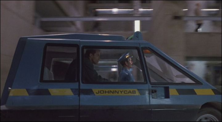 Side view of a small, boxy blue car, labelled "JOHNNYCAB" on a yellow and dark blue stripe, with the front pointed to the right.  Inside the car is Arnold Schwarzeneggar leaning forward, and there's a maniken "dressed" in blue as the driver.   In the background is brutalist architecture.
Frame grabbed from a scene from the 1990 movie Total Recall.