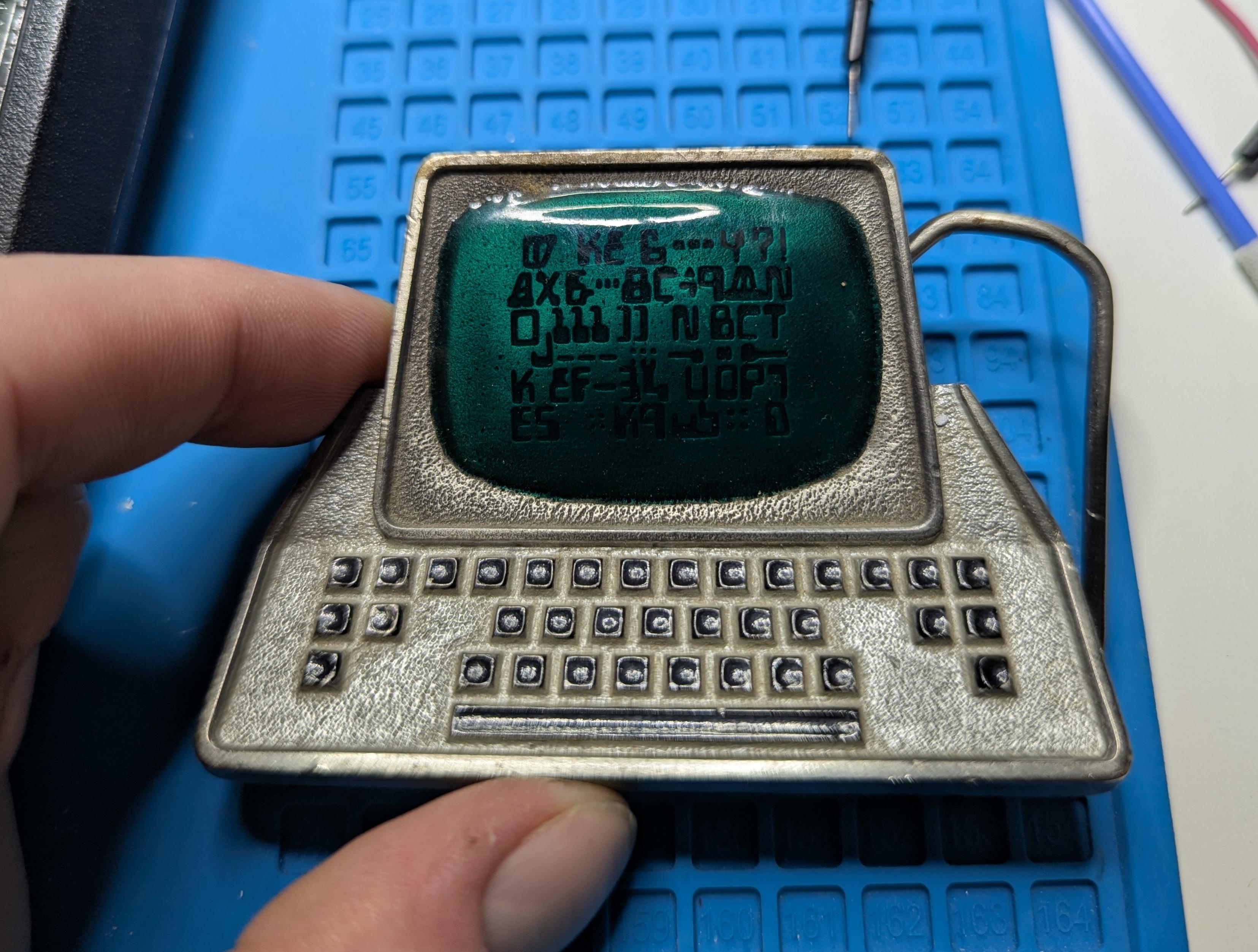 A comically oversized pewter belt buckle in the shape of a stylized 1970s green screen terminal.