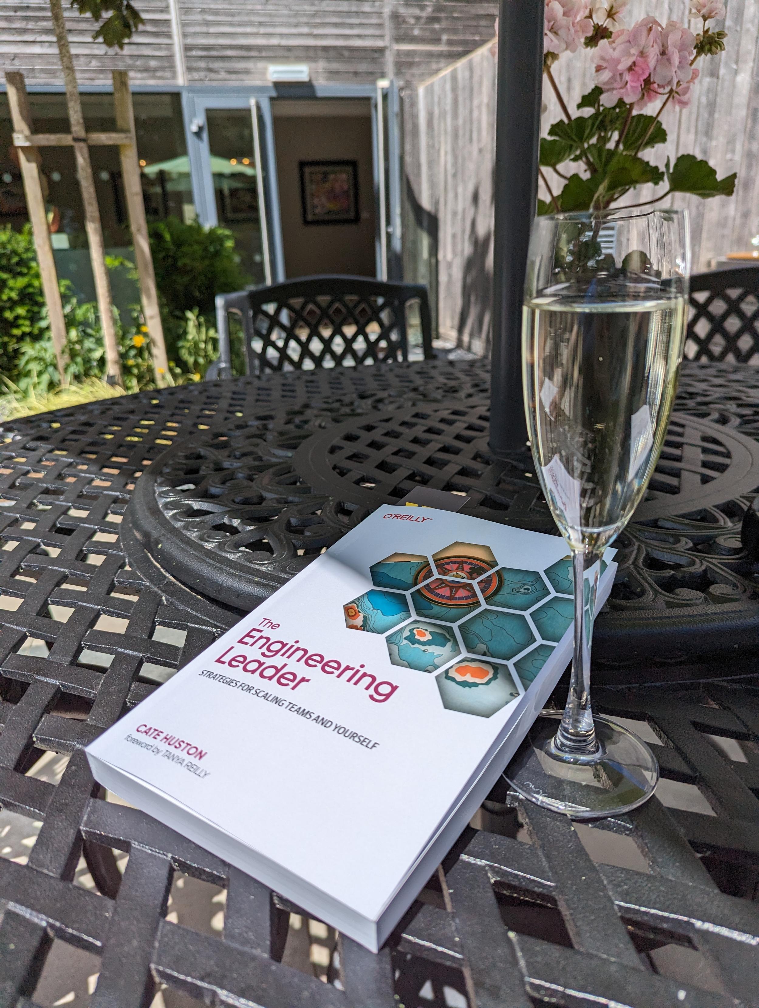 A picture of the engineering leader book next to a glass of fizz on a table. It's outside, there is sunlight and flowers