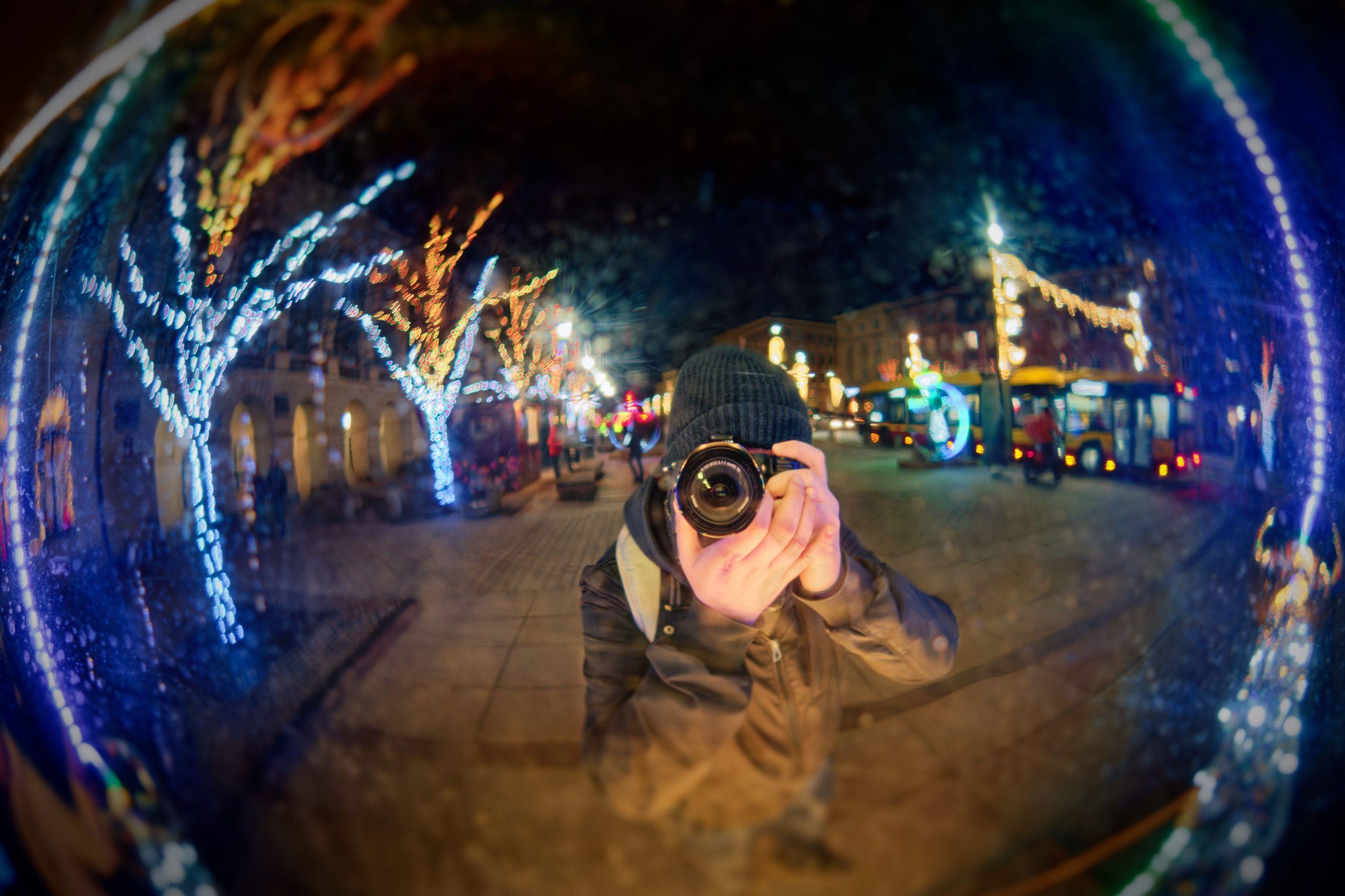 Me taking a selife with a camera against a glossy Christmas installation in Warsaw Old Town. Various Christmas lights and city nightscape visible in the background. February 2024.