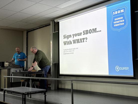 Photo of the stage, with two people looking down at a desk. There is a large screen showing an image with the text: Sign your SBOM… With WHAT?