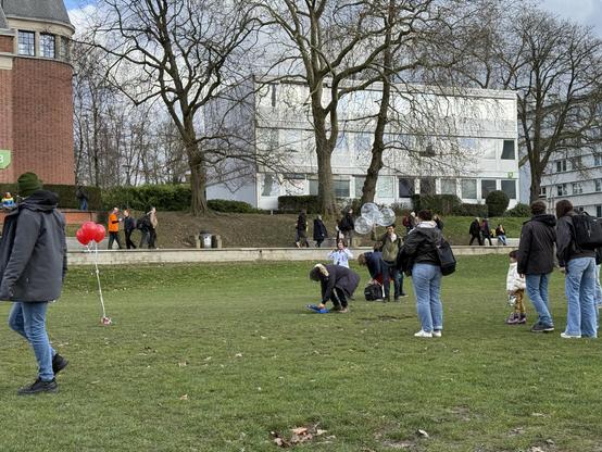 Photo of a grassy field with a few red balloons, and some people fiddling with something on the ground. 