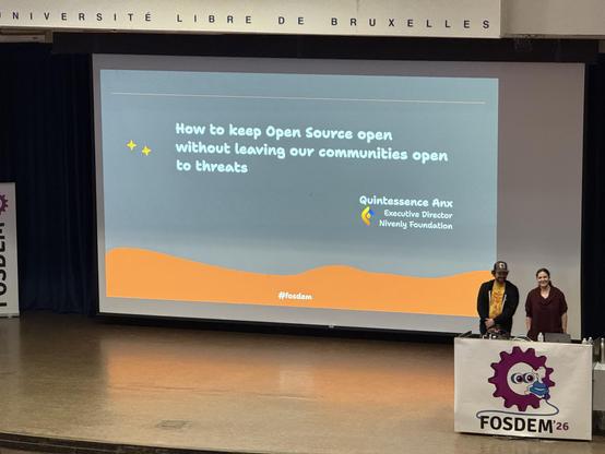 Photo of a stage with two people standing behind the podium. On the screen there is the text: How to keep open source open without leaving our communities open to threat