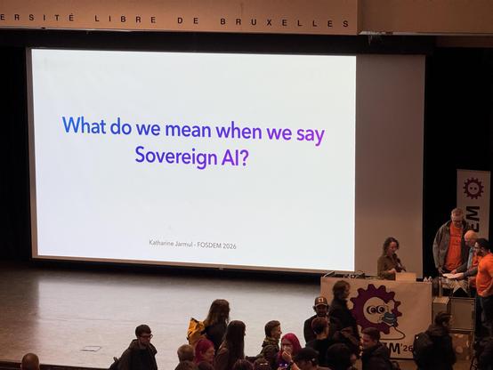 Photo of the stage with some people milling about. The text on the screen is: What do we mean when we say Sovereign AI