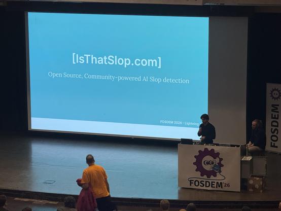 A dark stage with a person behind the podium. The screen says: IsThatSlop.com Open source. community-powered AI slop detection. 
