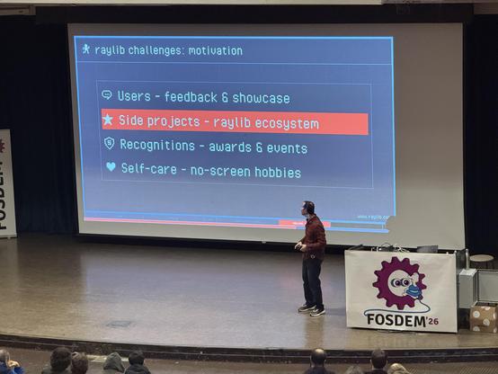 A person on stage looking up at a screen with the text:

Raylib challenges: motivation 
Users - feedback & showcase
Side projects- raylib ecosystem 
Recognitions - awards & events
Self-care - no-screen hobbies