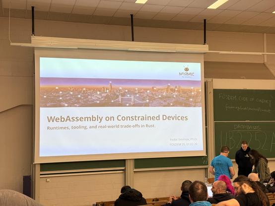 Photo of a few people on a low stage. There is a screen with the text: WebAssembly on constrained devices