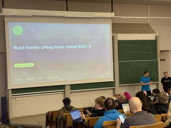 Photo of a stage with a screen saying Rust meets cheap bare-metal RISC-V