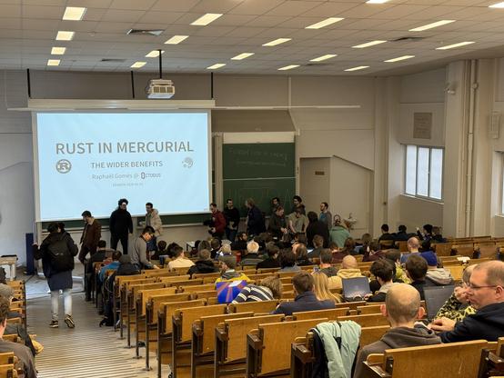 Photo from the back of a devroom with a slide showing on the screen with the text: Rust in Mercurial