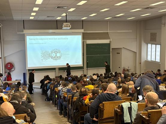 Photo from the back of a filled devroom showing a slide with the title: Taming Git complexity with Rust and Gitoxide
