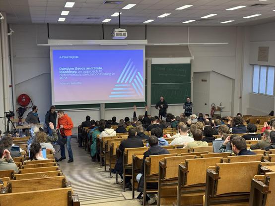 Photo from the back of a devroom showing a screen with a slide saying: Random Seeds and State Machines: an approach to deterministic simulation testing in Rust