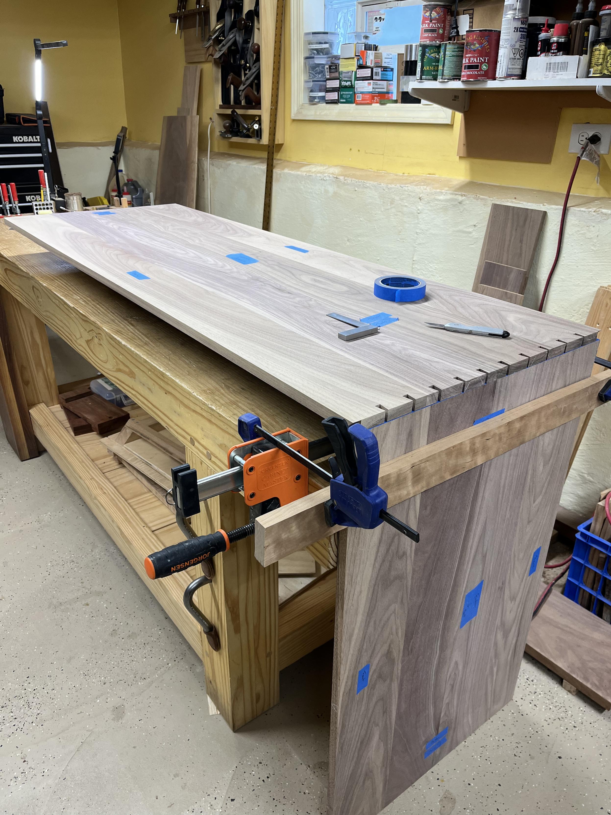 The long side piece of a cabinet case laying flat on top of a workbench. It's elevated about six inches off the workbench top. On the end of the workbench is another shorter piece that meets the end of the long piece at a 90 degree angle. The long piece is sitting on top of the end of the short piece so that dovetail marks can be transfered to the short pin board piece.