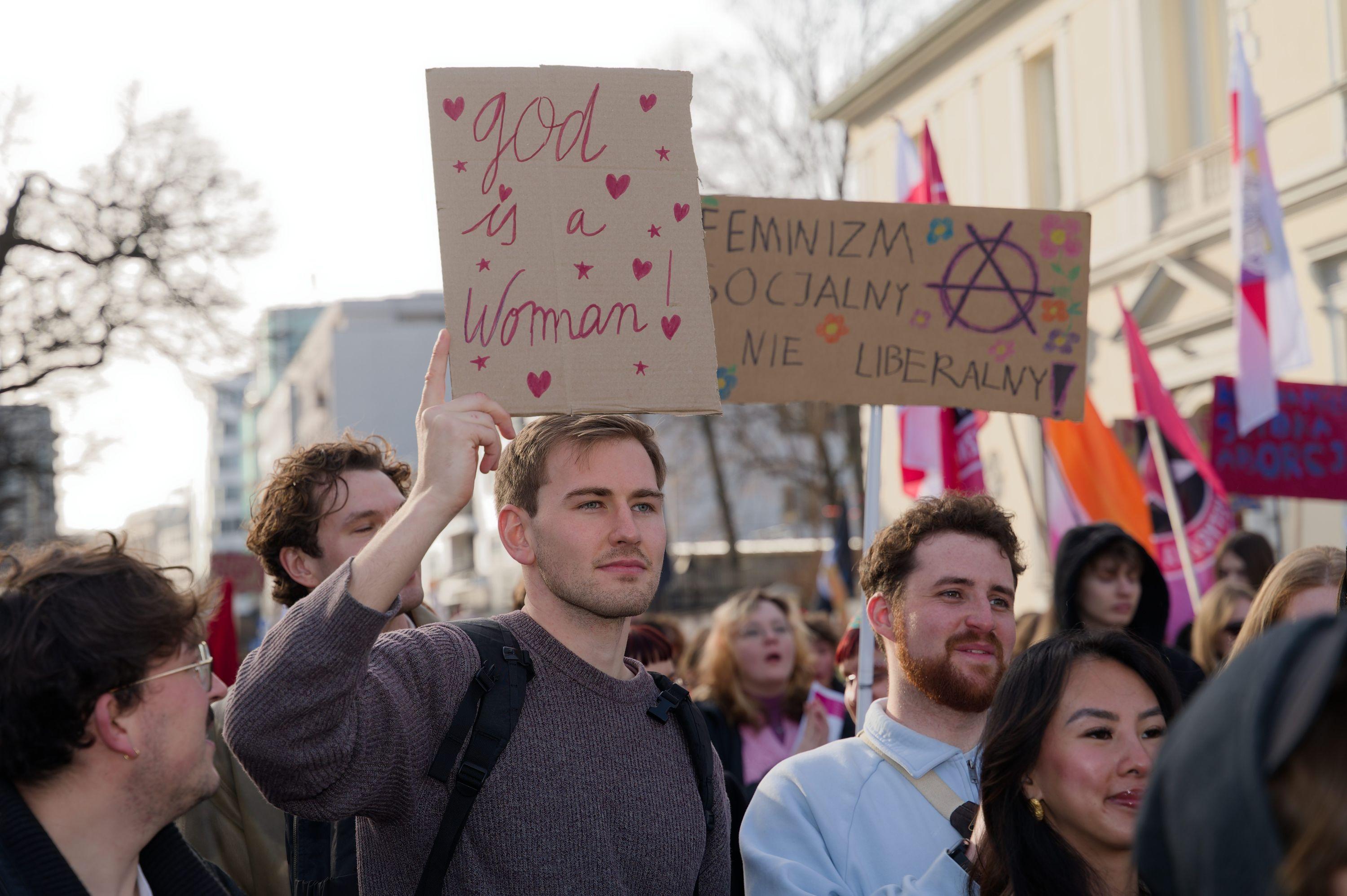 A group of men, one of them is holding a placard saying "god is a woman!"