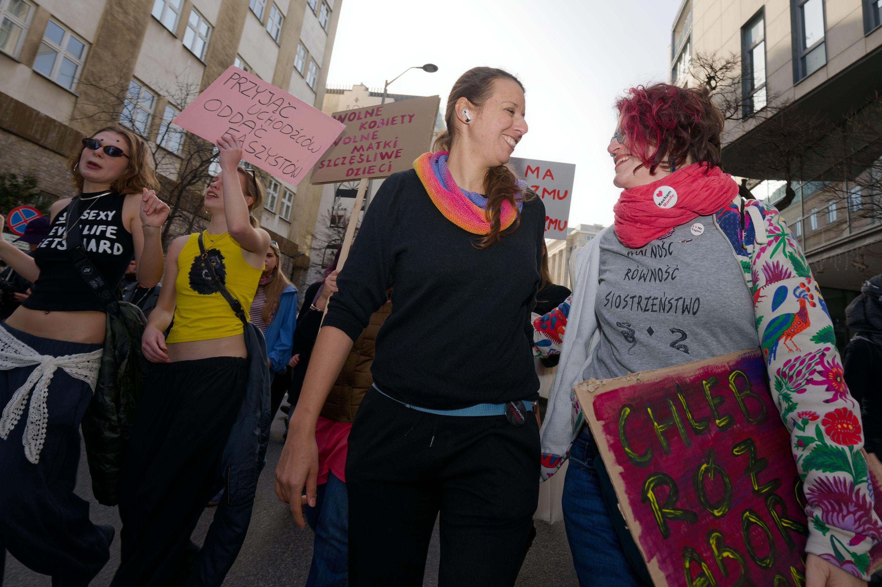 Two women looking at each other, smiling and exchanging friendly gestures while walking alongside a dancing crowd.
