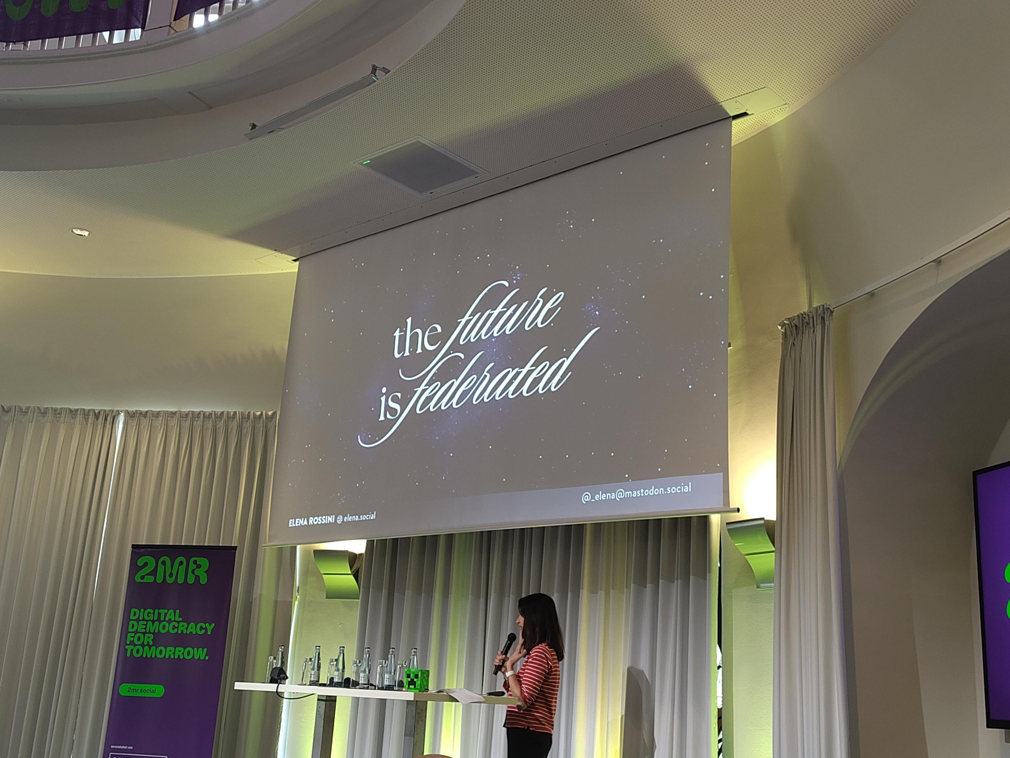 Photo of a person standing in front of a projector with the text "the future is federated"
