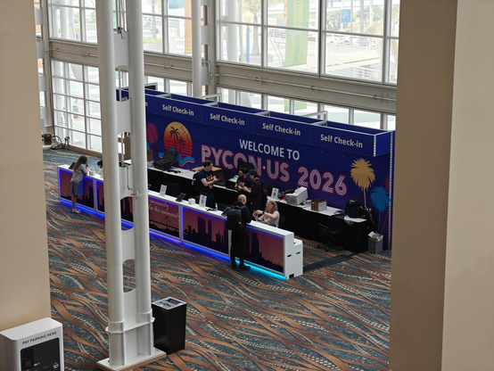 View from second floor down to the registration desk at PyCon. Several people behind the registration area. A big sign behind them says "Welcome to PyCon US 2026"