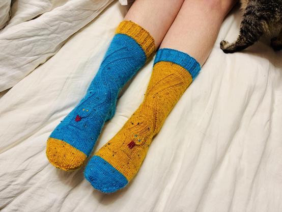 A pair of socks, one mostly blue with a yellow toe and cuff, and the other yellow with a blue toe and cuff. Each is knit with a snake formed by the texture of the fabric, and the snakes are sticking out their wee tongues. A cat’s back leg is visible in the top right corner of the photo.