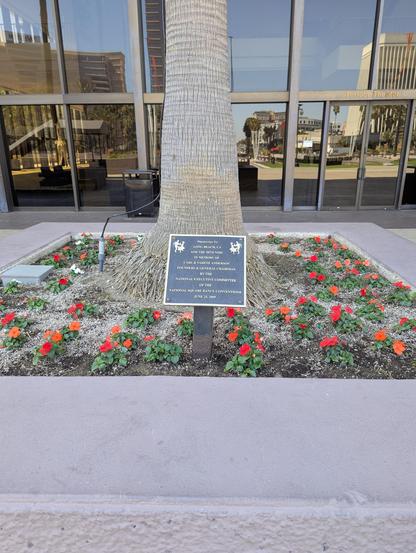A plaque surrounded by flowers at the base of the palm tree. in the upper corners, one couple per corner is depicted dancing. The text in the middle reads:
"Presented to
Long Beach, CA
And the 58th NSDC
In memory of
Carl & Varene Anderson
Founders & General Chairman
by the
Executive Committee
of the 
National Square Dance Convention®
June 25, 2009" 