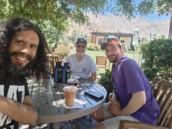 Three sentimental men sitting around a shaded table enjoying coffee and company.