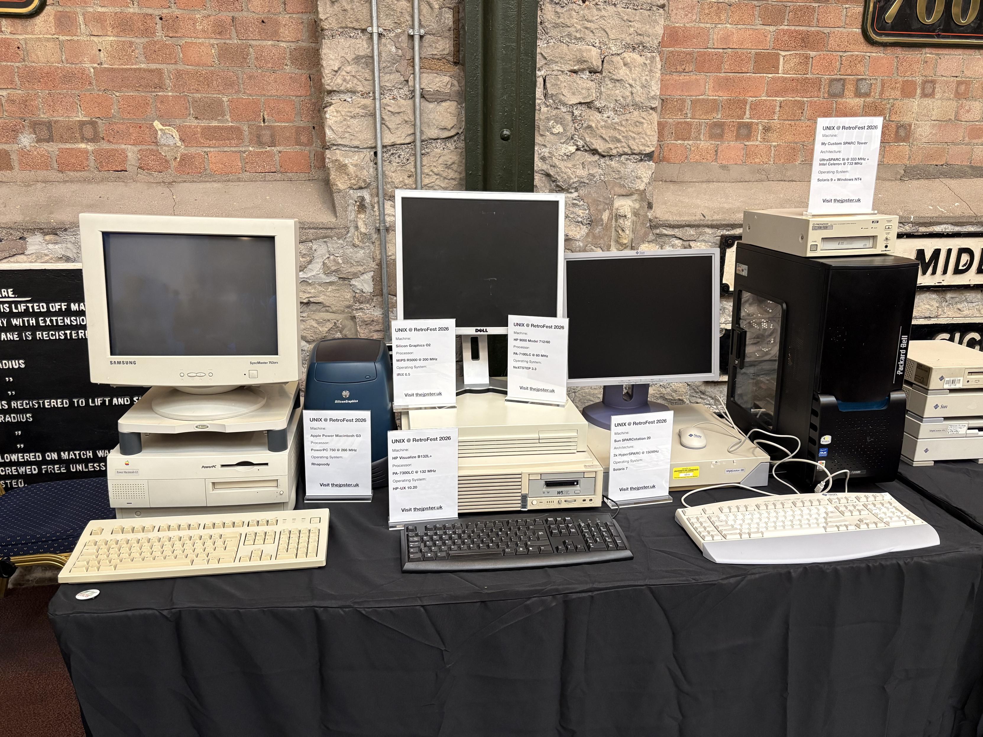 A table at an exhibition. It has a table cloth. It has six computers and three monitors on it.