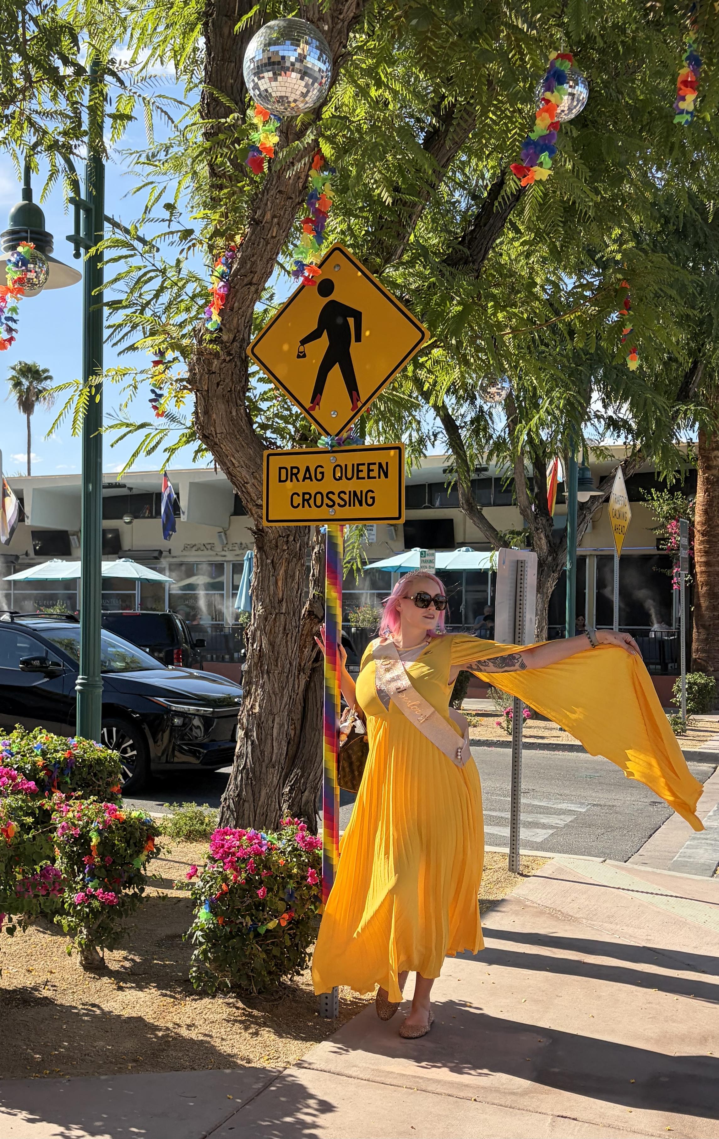 Pink haired woman in a yellow dress with giant sleeves standing next to a sign that says “drag queen crossing”