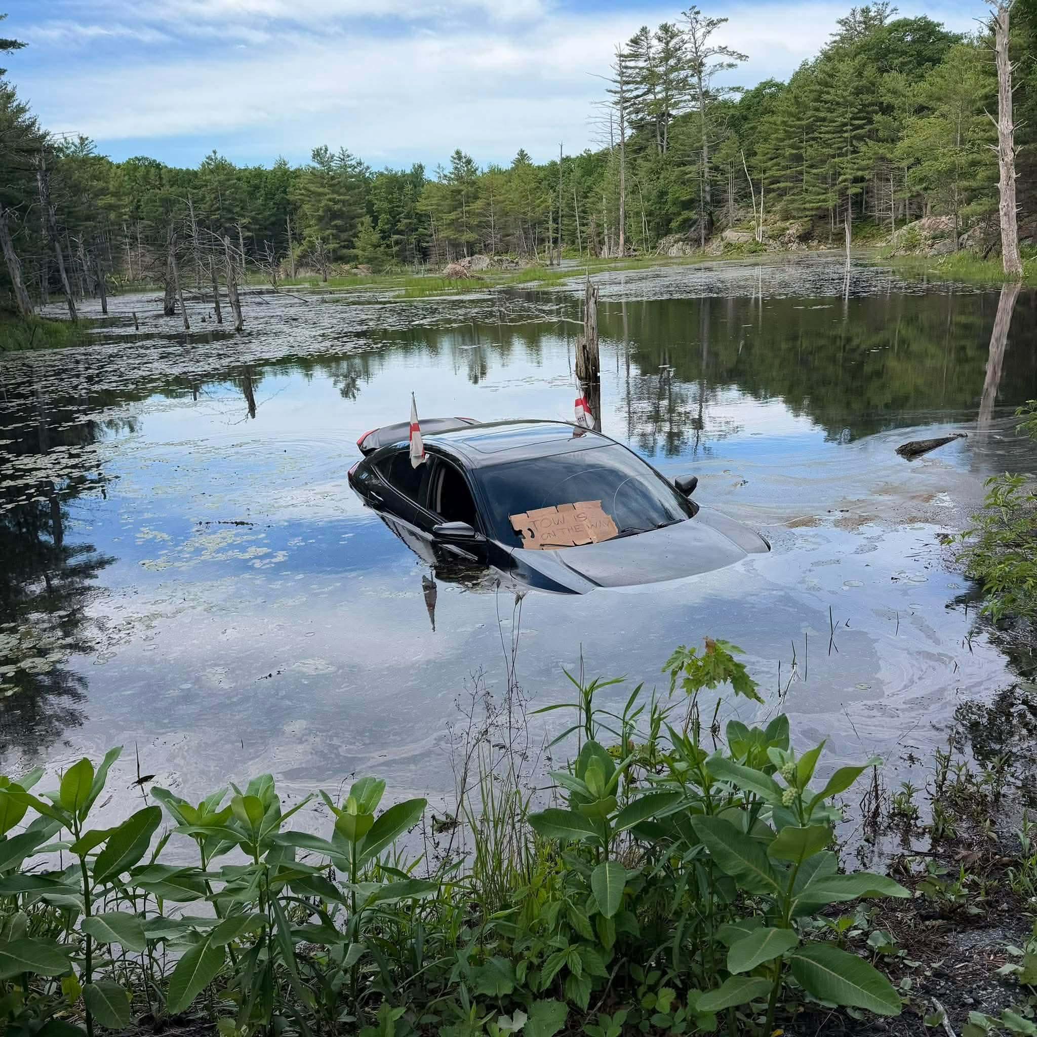 Photo of a marsh, with an open area of perfectly still water in the middle, and a green forest surrounding it.  A cloudy blue sky is reflected in the open water.  An automobile also sits in the water, submerged up to the level of the side mirrors.  In the front window of the car is a hastily-drawn cardboard sign that says “Tow is on the way”.
