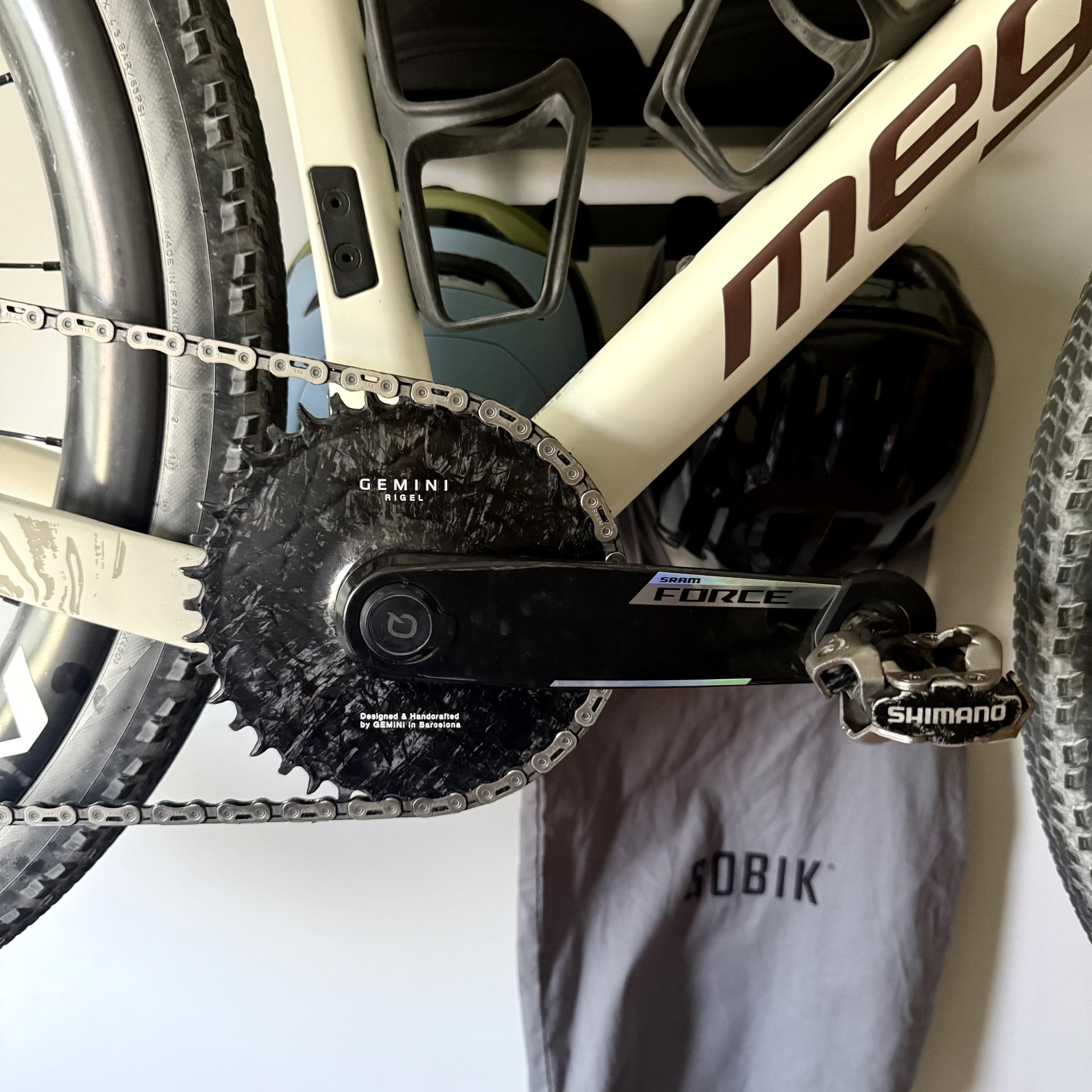 Alt-text:

Close-up of a gravel bike drivetrain showing a black carbon Gemini chainring, SRAM Force crank arm with integrated power meter, and a Shimano XT SPD pedal. The cream-colored bike frame has two bottle cages, with helmets and cycling gear visible in the background
