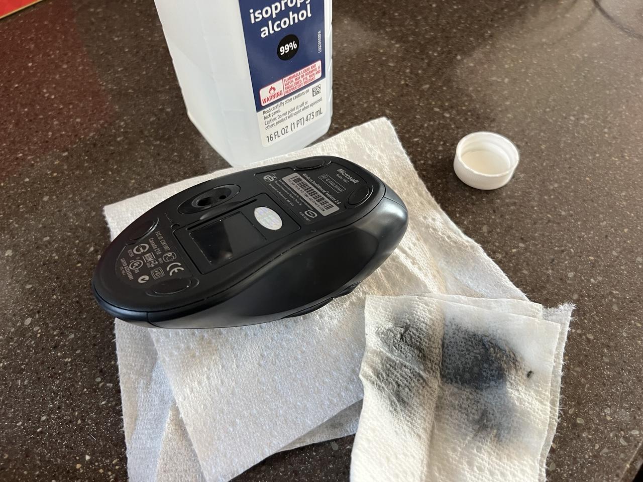 Microsoft IntelliMouse Explorer 2.0 mouse with a semi-shiny surface which was previously very sticky grippy coating. The mouse sits next to a bottle of isopropyl alcohol and a black-stained paper towel used to remove the coating.