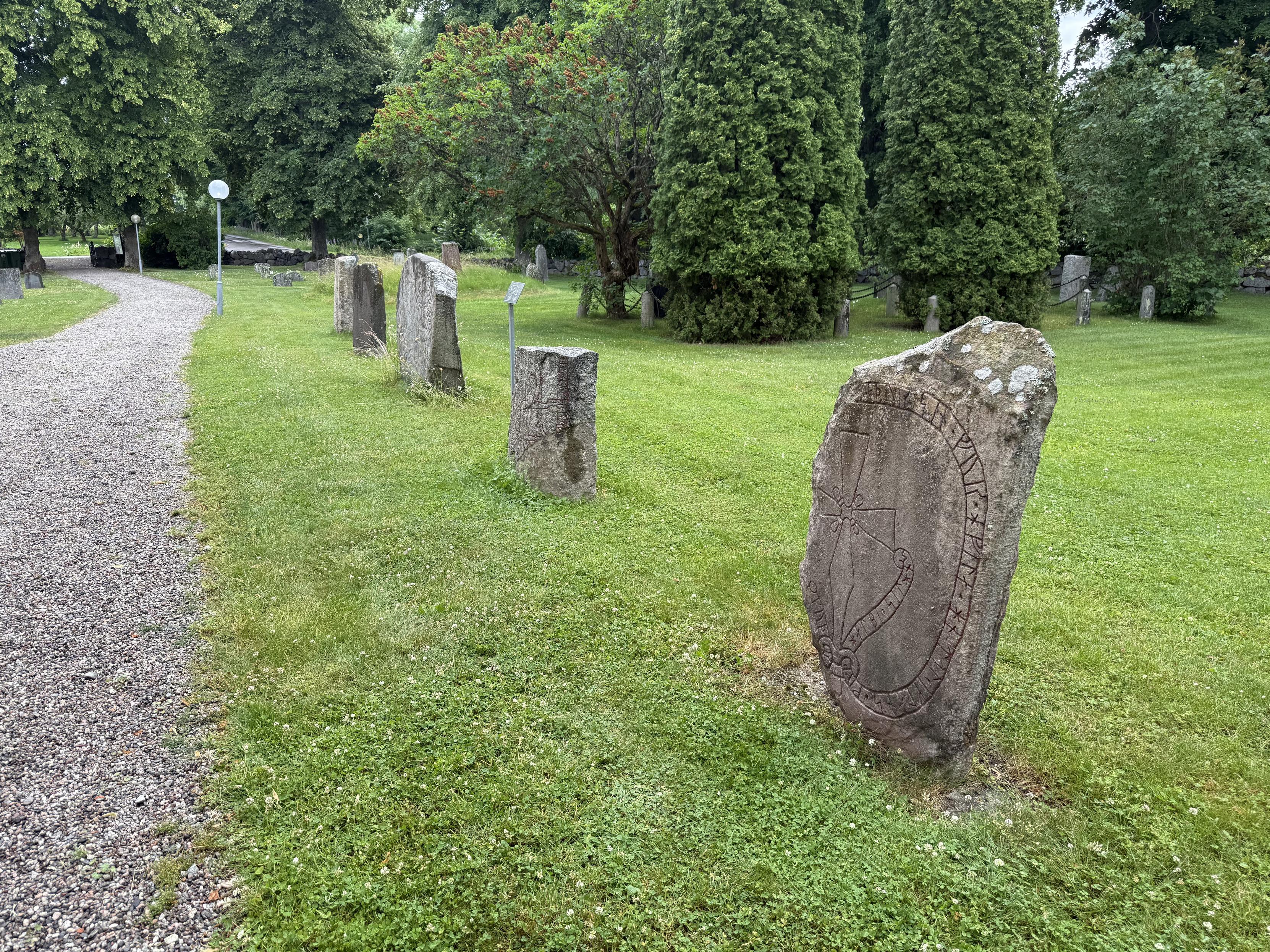Photo of five runestones standing next to each other next to a gravel road. There are some green trees in the background.
