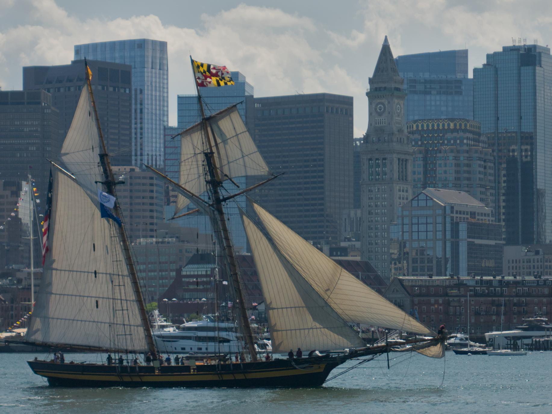 The schooner Pride of Baltimore sailing into Boston Harbor as part of Sail Boston 2026. The Custom House Tower and other parts of the downtown Boston skyline are visible in the background.