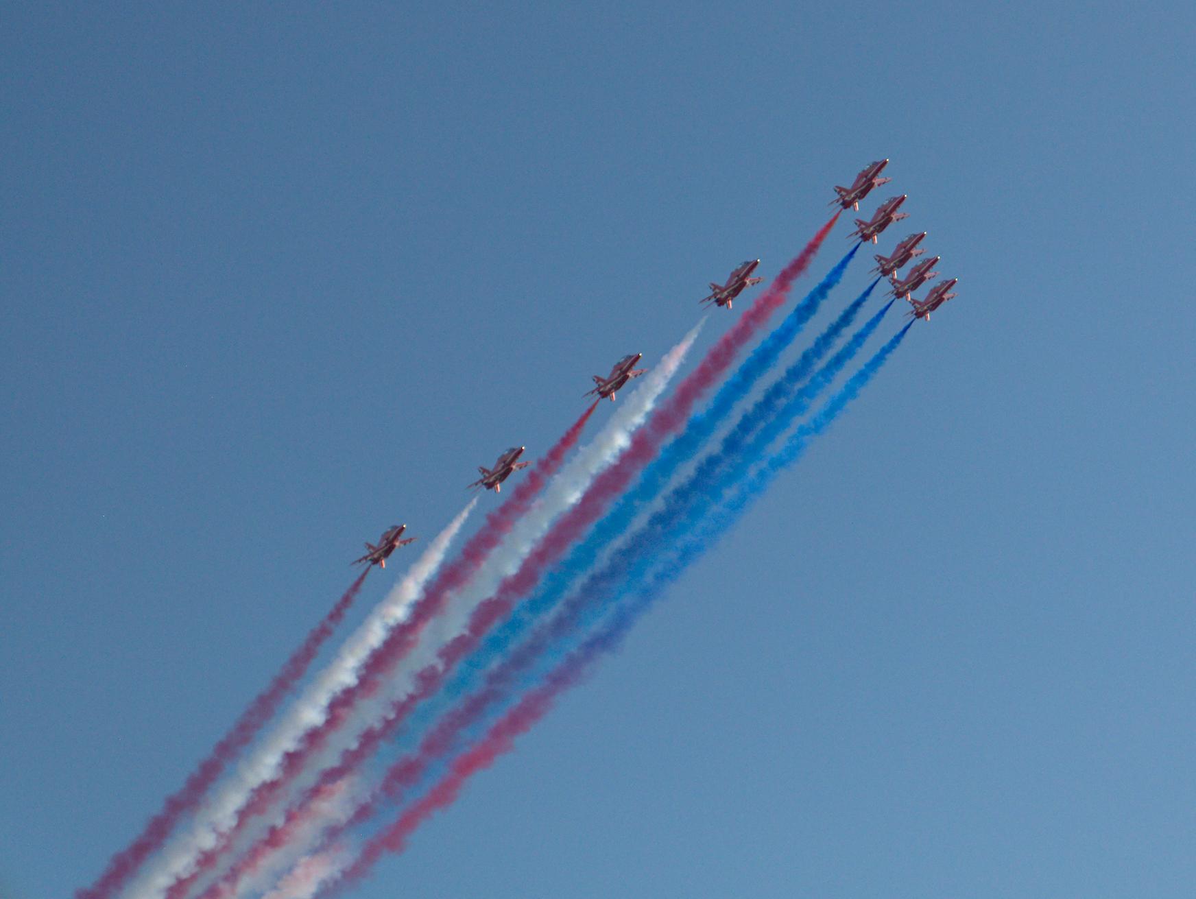 UK Royal Air Force "Red Arrows" flying in a chevron formation above the Boston Harbor, trailing red white and blue smoke evocative of the US flag. The plane of the formation is tilted slightly towards the viewer, giving a slight sense of vertigo