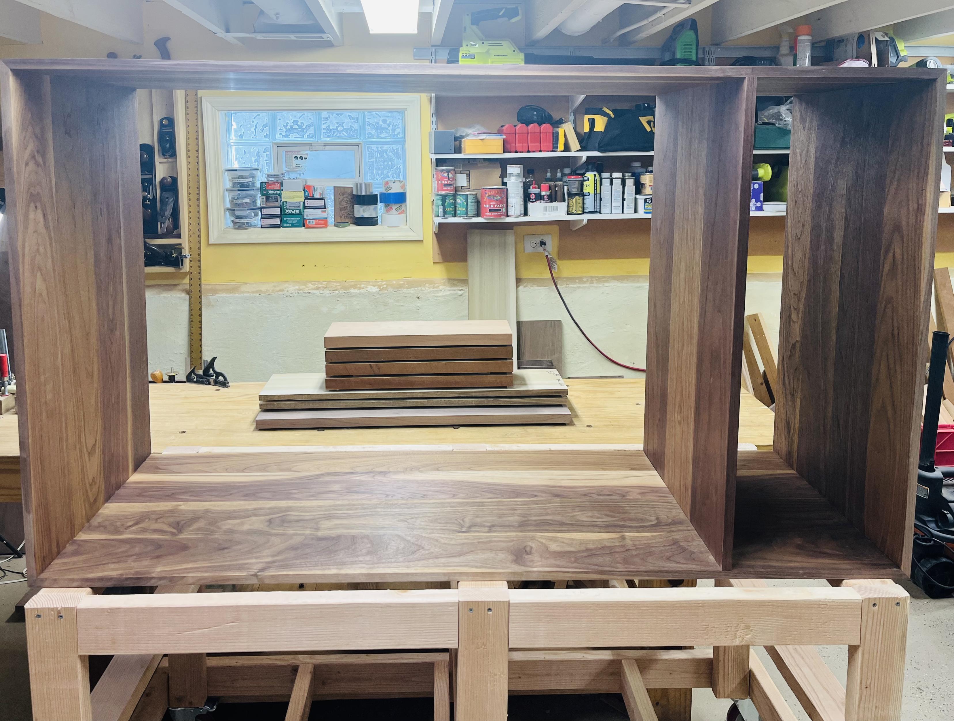 A large rectangular cabinet carcass laying on its side on top of a cart made of 2x4 wood. There is a vertical divider in the cabinet where a drawer will fit on the bottom and a larger section at the top. You can see through it where a stack of wood sits on a woodworking bench.
