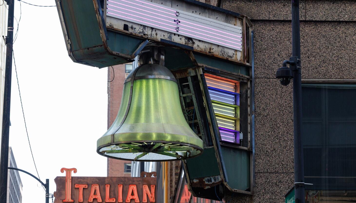 A bell-shaped object with green light emanating from it hangs from the frame of an old neon sign on the corner of a mid-century office building. The signs neon tubes are exposed and also give off light in various colors.