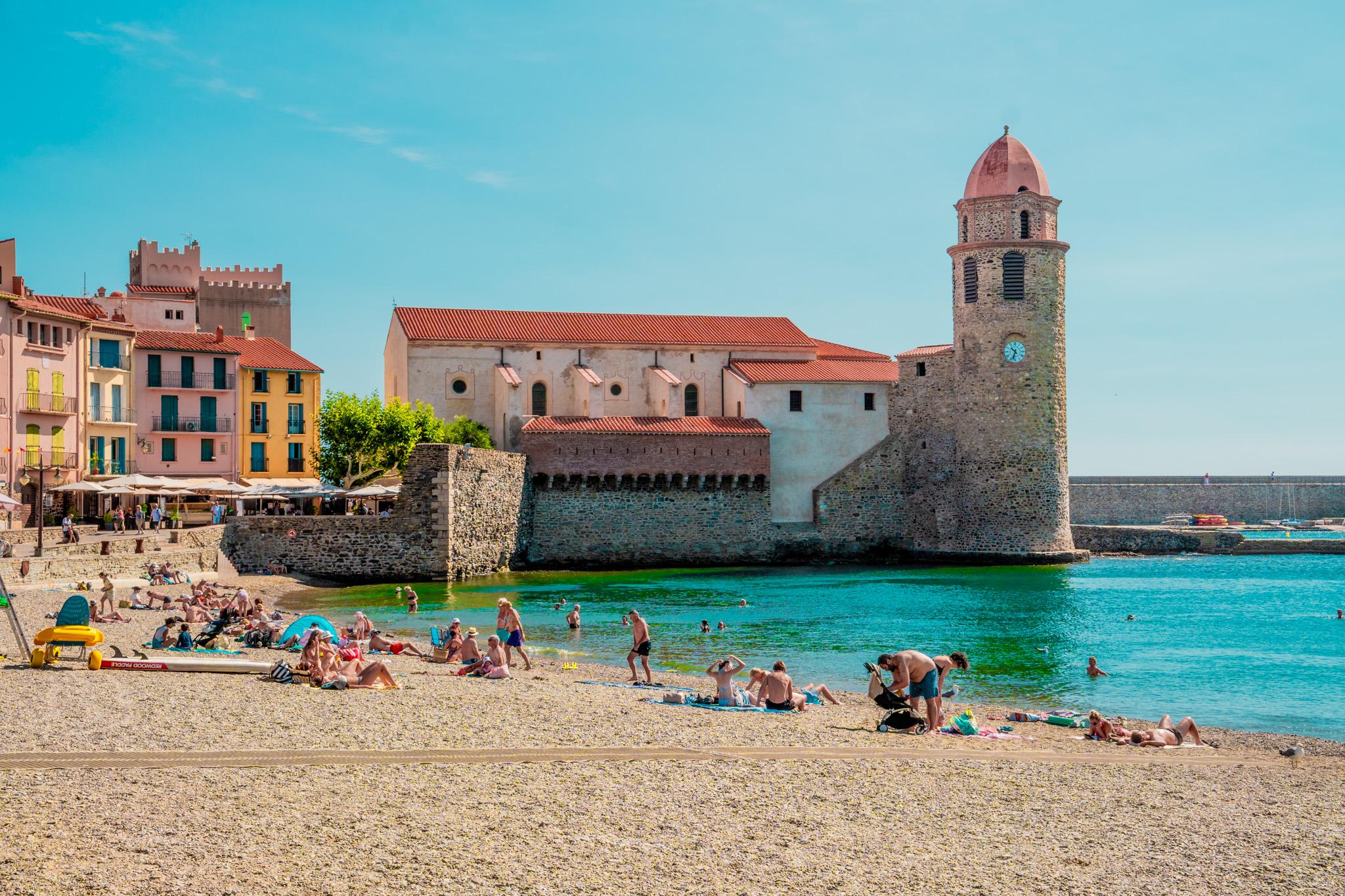 a beach in Collioure france in the foreground. People are sunbathing there. In the background you see an old tower-castle like structure. The sky is very blue.