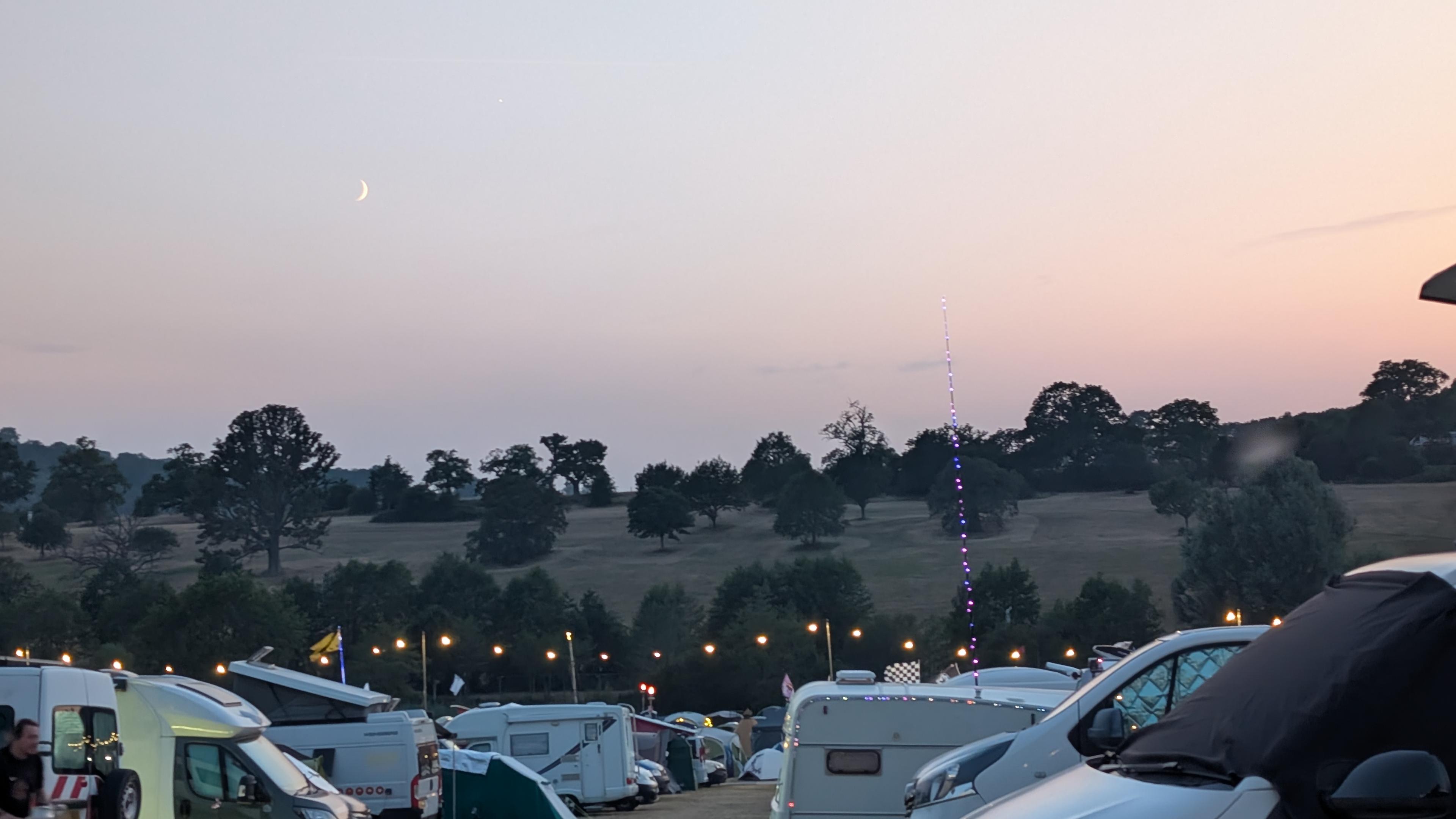 a sliver of a moon at dusk. there are camper vans and caravans in the foreground and a hill with trees in the distance