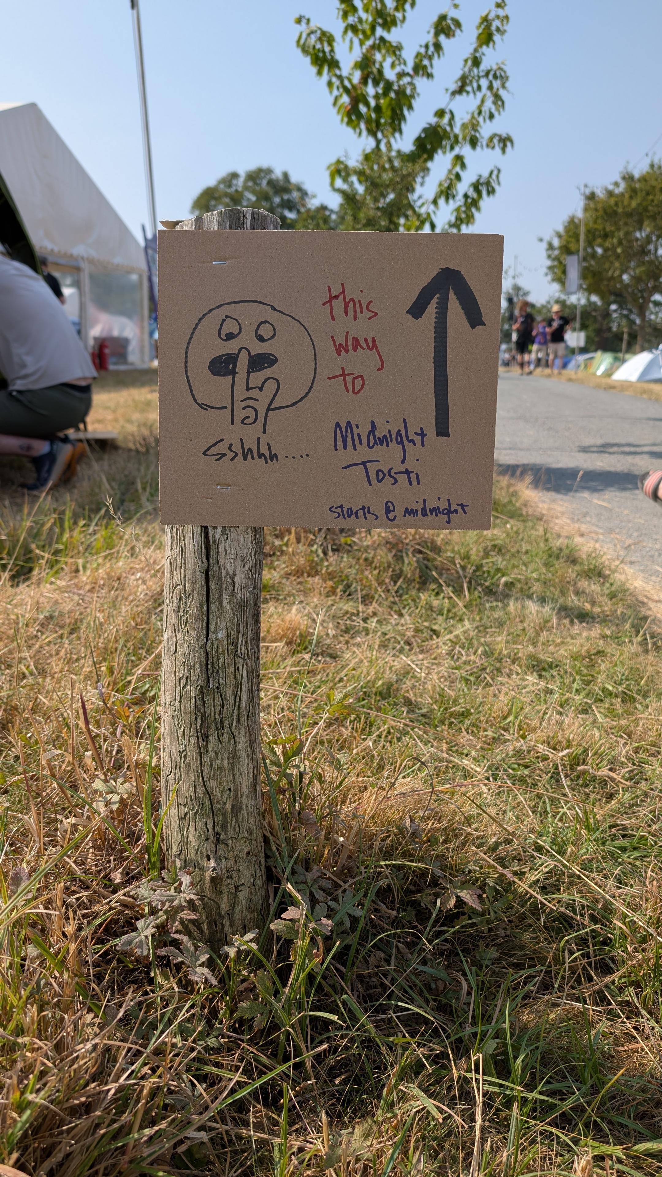 a small brown cardboard sign with a hand drawn 🤫 emoji. the test reads This Way to Midnight Tosti
