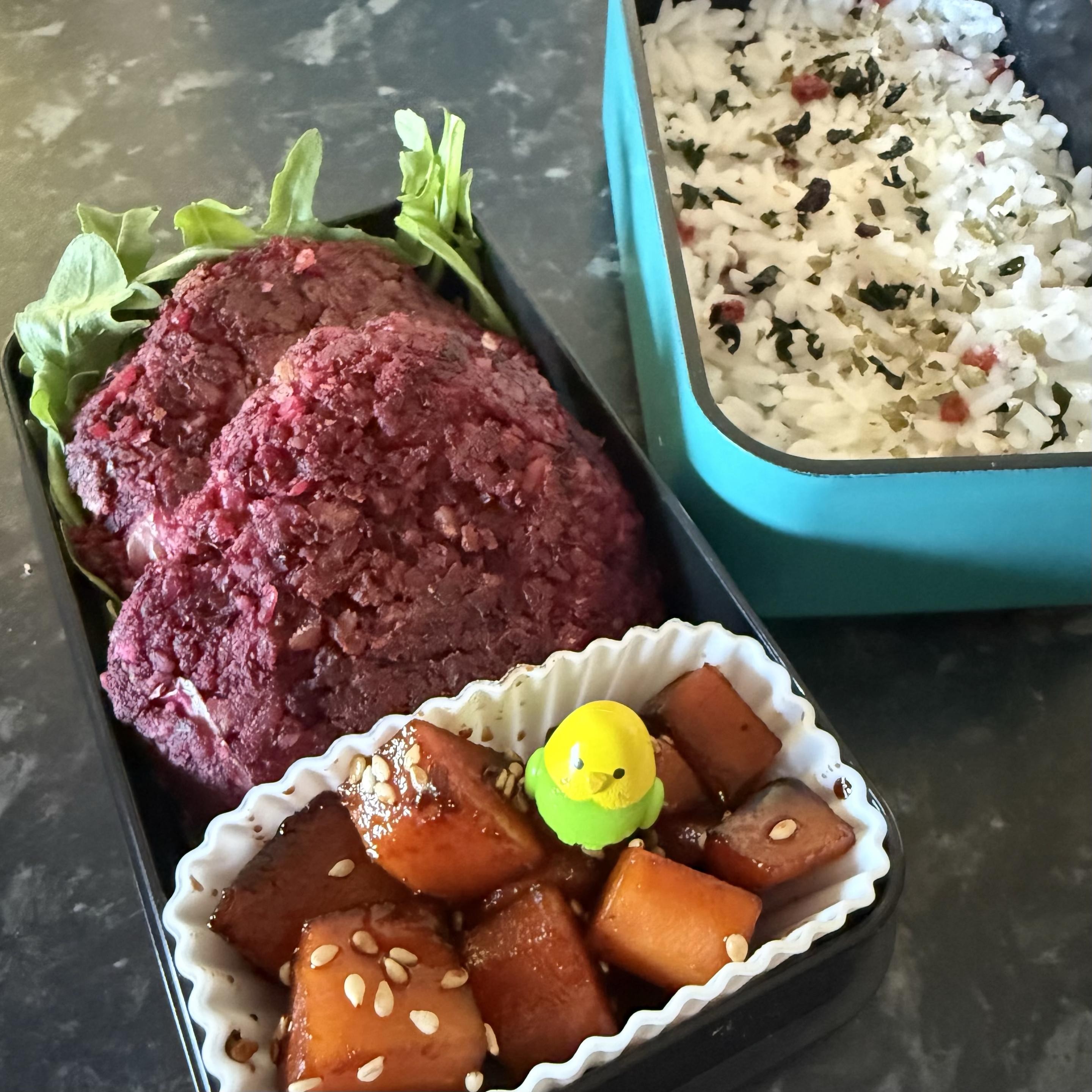A bento box featuring 2 bright beetroot burgers surrounded by leafy greens and a cupcake case with carrots in a dark brown sauce topped with sesame seeds and a small decorative budgie. On the right there is a separate compartment of seasoned white rice.