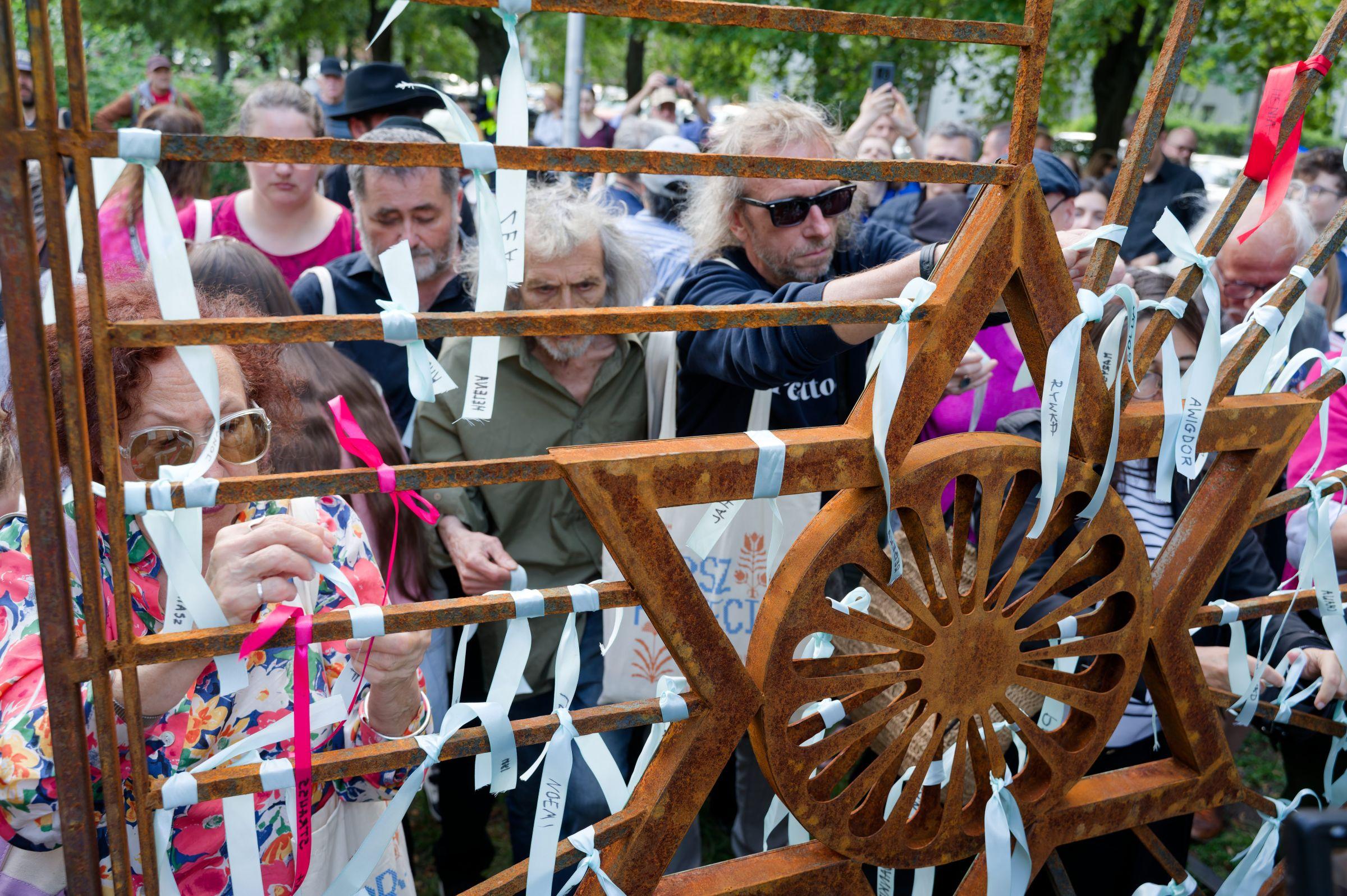 Crowd of people tying Ribbons of Remembrance featuring names of Holocaust victims to the art installation made by Zuzanna Hertzberg at Rachel Auerbach square.