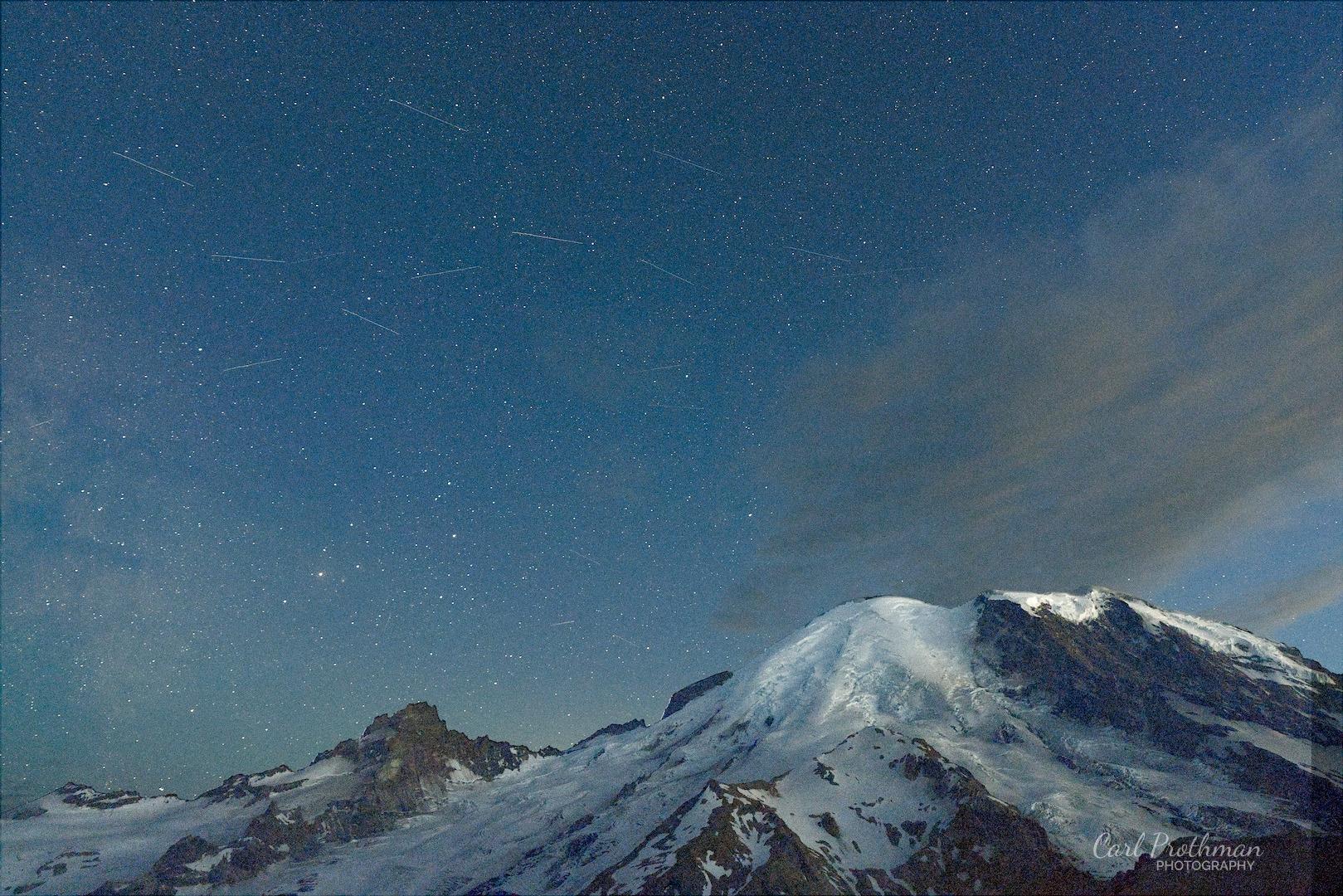 Mount Rainier rises beneath a wide night sky filled with countless stars. Snowfields and glaciers remain visible across the mountain’s upper slopes, while the lower ridges fade into darkness. Thin clouds drift across the right side of the sky, adding softness and texture among the sharper points of starlight. Several faint satellite trails create narrow lines across the stars. The scene feels vast, quiet, and peaceful.