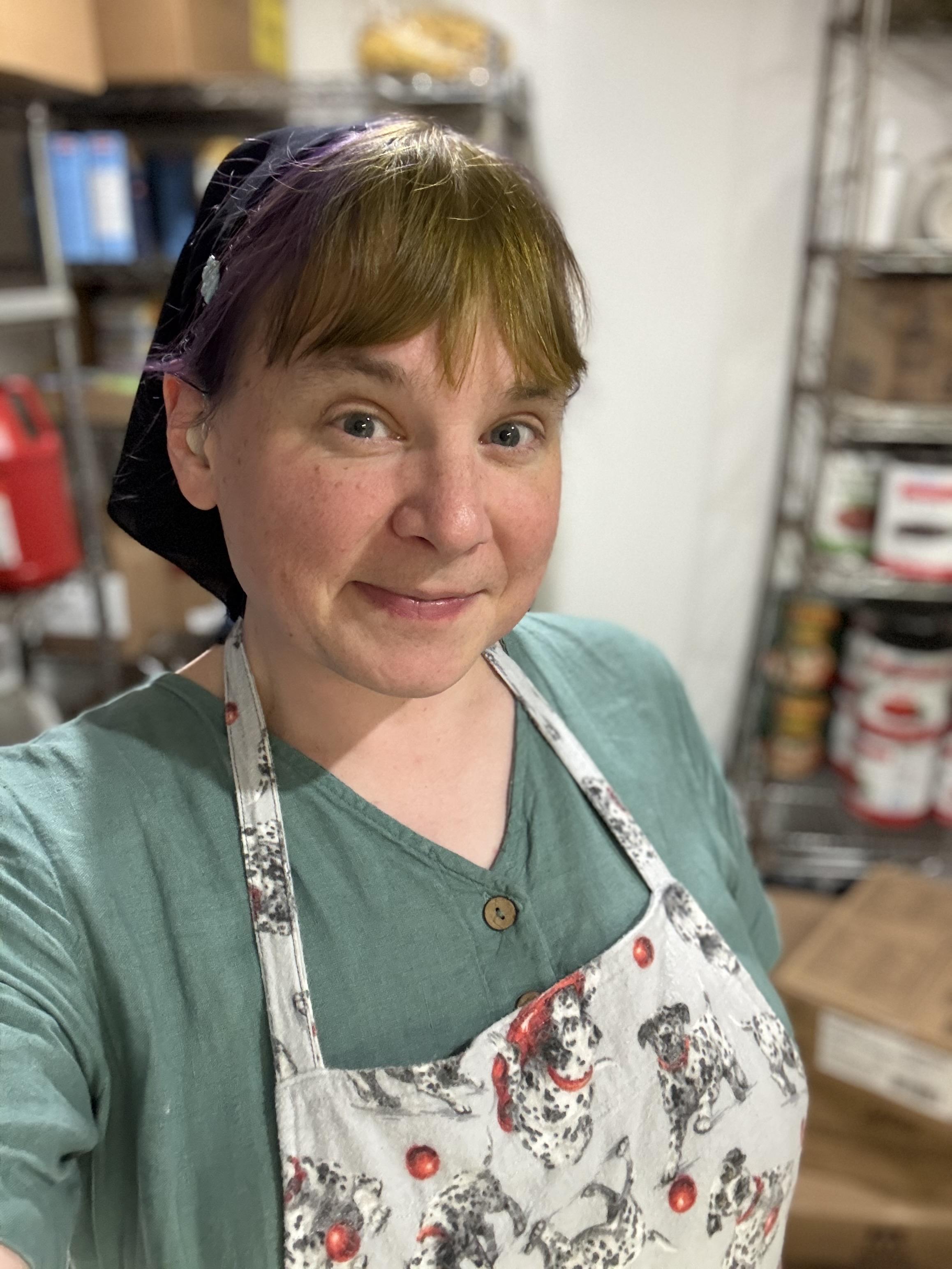 Sen, in a green dress looking at the camera wearing a Dalmatian-themed apron standing in a pantry, with the background too out of focus to make out specific items but with many things on wire shelving.