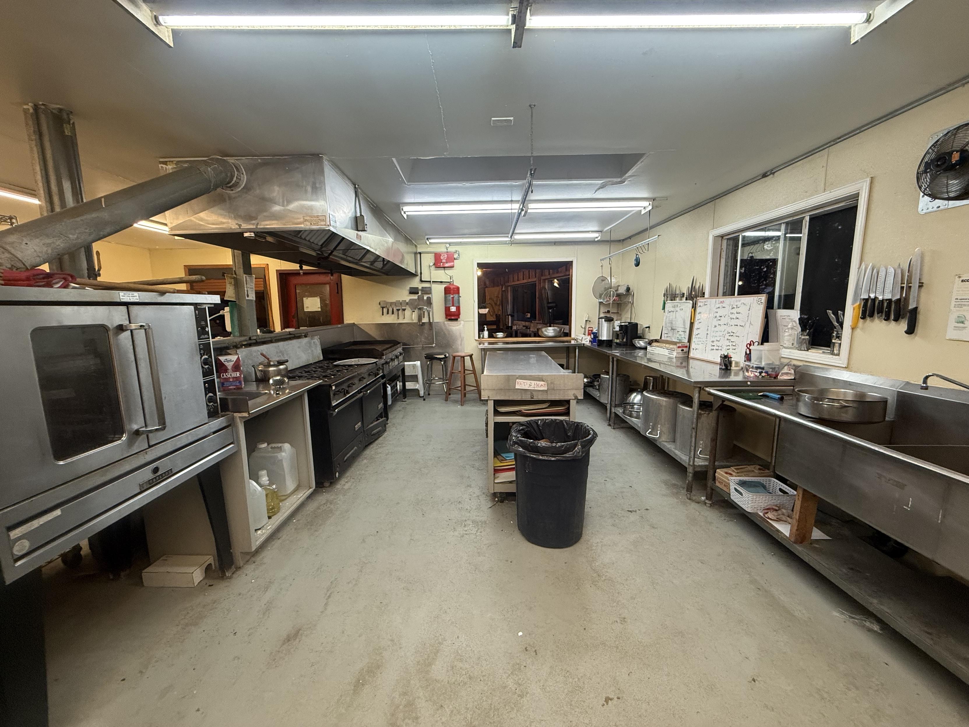 A commercial kitchen, on the left side from close to near a convection oven, a small sink area, 12 burners worth of gas stove, and a flat top grill. The right side has a two compartment sink, a long counter with whiteboards on it, and a window behind. An island with a steel countertop is in the centre, with a window at the far end looking out into a camp dining hall